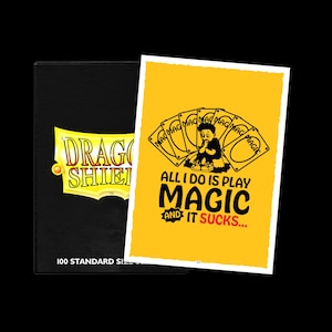 Op de afbeelding: Een zwarte kaartenopbergdoos met de tekst "DRAGON SHIELD" in geel en wit. Daarnaast een gele kaart met een cartoonafbeelding en de tekst "ALL I DO IS PLAY MAGIC AND IT SUCKS..."