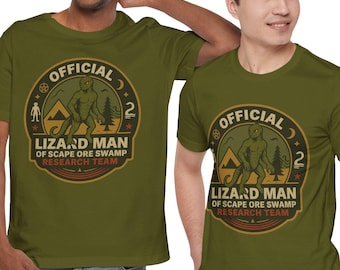 May include: Two olive green t-shirts with a circular graphic that reads "OFFICIAL LIZARD MAN OF SCAPE ORE SWAMP RESEARCH TEAM." The graphic features a lizard-like creature, trees, and a mountain. The shirts are worn by two people.