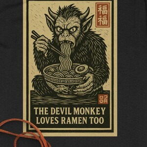 May include: A black and tan graphic featuring a horned monkey eating ramen with chopsticks. The text reads "THE DEVIL MONKEY LOVES RAMEN TOO" in a bold font. Japanese characters are in the top corners.