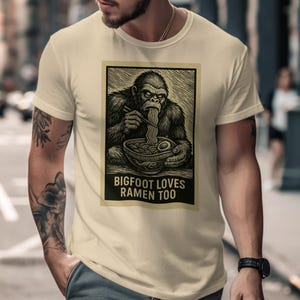 May include: Cream-colored t-shirt featuring a black and white illustration of Bigfoot eating ramen noodles with chopsticks. The text "BIGFOOT LOVES RAMEN TOO" is below the image. The shirt is short-sleeved.