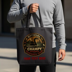 以下が含まれることがあります： 「OFFICIAL CHAMPY RESEARCH TEAM」の文字が入った円形デザインが特徴の黒いトートバッグ。デザインには、生き物、木、星が描かれた湖の風景が含まれています。「YOUR TEXT HERE」の文字がデザインの下にあります。