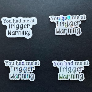 May include: Four stickers with the phrase "You had me at Trigger Warning" in a black font. The stickers have a holographic, iridescent effect, with a white border. The stickers are on a black background.