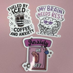 May include: Three stickers with humorous sayings. The first sticker features a skeleton hand holding a glass of iced coffee with the text "Fueled by Iced Coffee and Anxiety". The second sticker features a cartoon brain wearing sunglasses and relaxing in a chair with the text "My Brain Needs Rest". The third sticker features a light switch with the text "Anxiety On Off" with the switch flipped to the "Off" position.