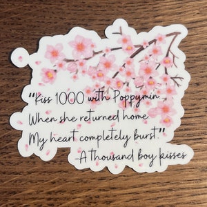 以下が含まれることがあります： 白いステッカー。ピンク色の桜の枝とテキストが特徴です。「Kiss 1000 with Poppymin. When she returned home. My heart completely burst!」-A thousand boy kisses. の引用句が書かれています。