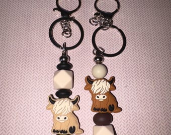May include: Two keychains featuring Highland cow charms. Each keychain has a silver-tone ring and clasp, with a series of beads in black, white, and brown. The cow charms are tan and brown with white fur.
