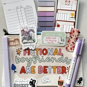 Puede incluir: Una bolsa de lona blanca con el texto "Fictional boyfriends are better" en letras de colores. La bolsa está decorada con varias pegatinas, incluyendo un dragón, un fantasma, un libro, un jugador de hockey y una taza de café. La bolsa también tiene un resaltador morado y un bolígrafo.