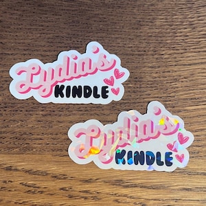 May include: Two stickers with the text "Lydia's Kindle" in pink and black lettering. The top sticker has a pink background and the bottom sticker has a holographic background.