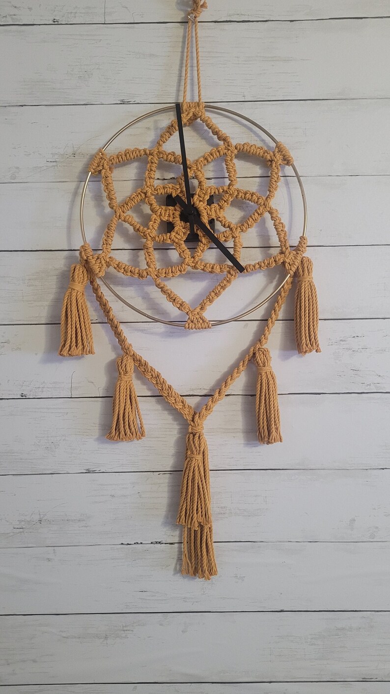 May include: A macrame clock with a gold metal ring and tan tassels hanging from the bottom. The clock is hanging on a white wood wall.