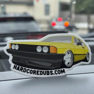May include: A die-cut sticker featuring a yellow car with black accents and the text "HARDCOREDUBS.COM". The car has a sporty design with white wheels and is set against a blurred background.