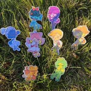 May include: A collection of eight die-cut stickers featuring stylized cartoon characters in various colors including blue, pink, yellow, and green. Each sticker has a holographic finish that reflects light, showcasing distinct character designs.
