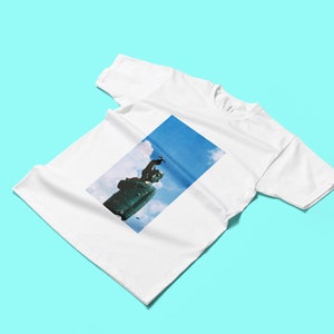 May include: White t-shirt featuring a rectangular image of a statue against a blue sky with clouds. The statue is dark green and appears to be made of metal. The t-shirt is made of a soft, white fabric.