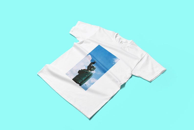 May include: White t-shirt featuring a rectangular image of a statue against a blue sky with clouds. The statue is dark green and appears to be made of metal. The t-shirt is made of a soft, white fabric.