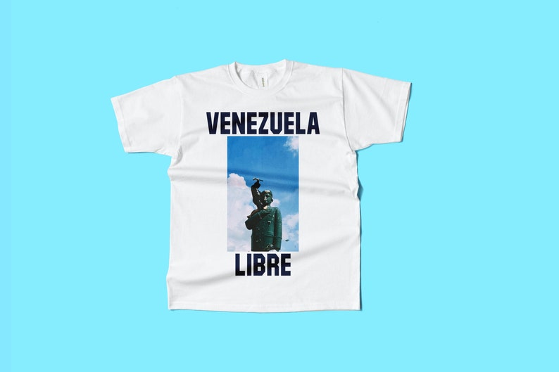 May include: White t-shirt with the words "VENEZUELA LIBRE" in navy blue. The shirt features a rectangular image of a statue against a blue sky background. The shirt is made of a soft, comfortable material.