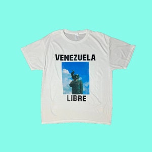 May include: White t-shirt with the words "VENEZUELA LIBRE" in black block letters. The shirt features a graphic of a statue against a blue sky background.