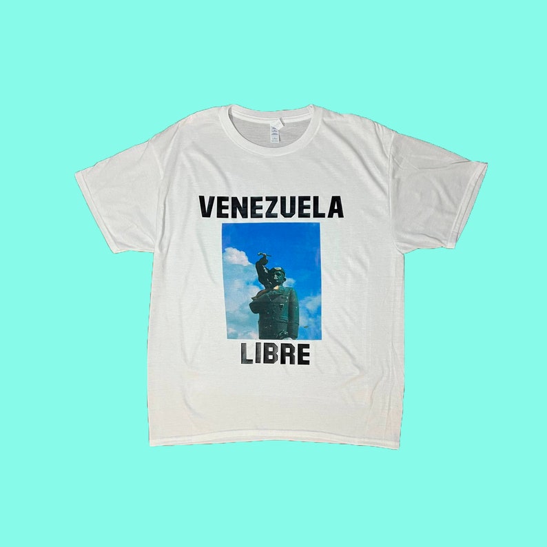 May include: White t-shirt with the words "VENEZUELA LIBRE" in black block letters. The shirt features a graphic of a statue against a blue sky background.