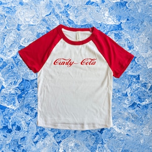May include: White and red raglan t-shirt with the text "Cunty-Cola" in red script across the front. The shirt has a white body and red sleeves. The background is a blue and white ice texture.