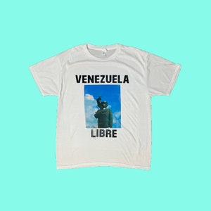 May include: White t-shirt with the words "VENEZUELA LIBRE" in black block letters. A rectangular image of a statue against a blue sky is centered below the word "VENEZUELA". The shirt is made of a soft material.