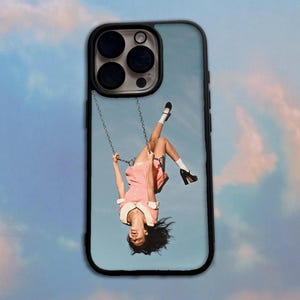 May include: A black phone case featuring a graphic print of a person in a pink dress with a white collar hanging upside down from a swing against a light blue sky background. The person wears white socks and black shoes while swinging in the air.