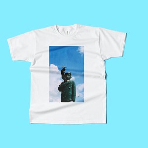 May include: White t-shirt with a rectangular graphic of a statue against a blue sky background. The statue is dark green and has a bird on its head. The t-shirt is displayed on a light blue background.