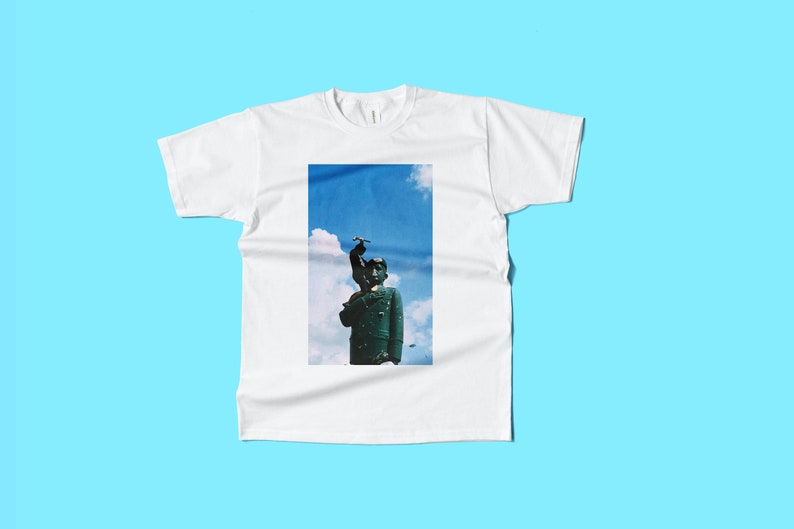 May include: White t-shirt with a rectangular graphic of a statue against a blue sky background. The statue is dark green and has a bird on its head. The t-shirt is displayed on a light blue background.