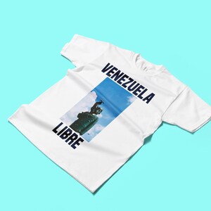 May include: White t-shirt with the words "VENEZUELA LIBRE" in navy blue. The shirt features a rectangular image of a statue against a blue sky. The t-shirt is displayed on a light blue background.