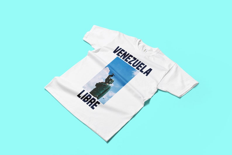 May include: White t-shirt with the words "VENEZUELA LIBRE" in navy blue. The shirt features a rectangular image of a statue against a blue sky. The t-shirt is displayed on a light blue background.