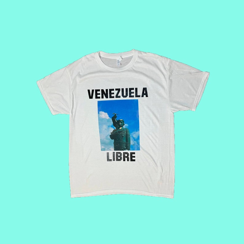 May include: White t-shirt featuring the words "VENEZUELA LIBRE" in black block letters above and below a rectangular image. The image depicts a statue against a blue sky background.