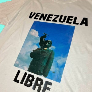 May include: White t-shirt with the words "VENEZUELA LIBRE" in bold black letters. The shirt features a graphic of a statue against a blue sky background.