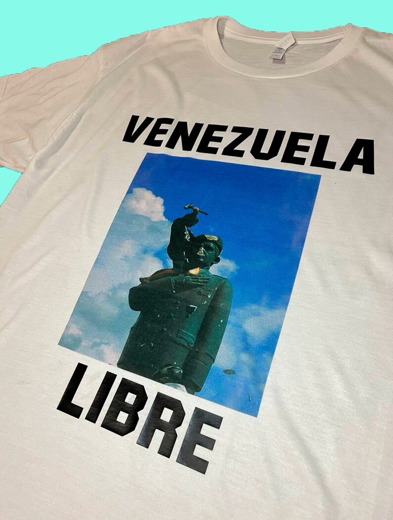 May include: White t-shirt with the words "VENEZUELA LIBRE" in bold black letters. The shirt features a graphic of a statue against a blue sky background.