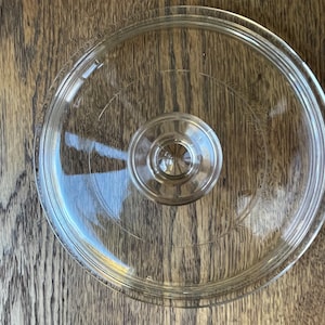 May include: A clear glass lid with a circular design and a central knob. The lid has a slightly raised rim and etched details. The glass is transparent, allowing a view of the wooden surface beneath. A kitchenware item.