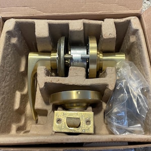 May include: A gold door handle and lock set in a cardboard box with foam packaging. The handle has a lever design and the lock has a round shape.