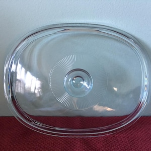 May include: Clear glass oval-shaped lid with a raised knob in the center. The lid has a textured pattern around the knob.