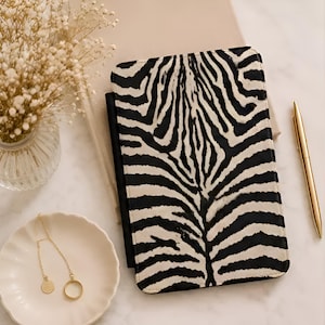 May include: A rectangular notebook cover featuring a black and white zebra stripe pattern. The cover is positioned vertically and includes a black pen tucked into the side. The design displays a symmetrical arrangement of stripes across the front surface.