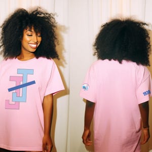 Peut inclure: T-shirt rose clair avec un motif g&eacute;om&eacute;trique bleu clair et rose. Le t-shirt a des manches courtes et un col rond. Le dos du t-shirt porte un petit &eacute;cusson et le chiffre "1938" imprim&eacute; en bleu.