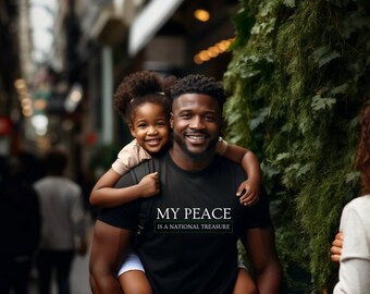 May include: A Black man is carrying his daughter on his back. He is smiling and wearing a black t-shirt with the text "My Peace is a National Treasure". The girl is smiling and wearing a light pink shirt and denim shorts.