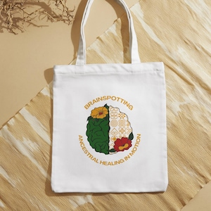 Può includere: Borsa tote bianca con una grafica di un cervello con fiori e il testo "Brainspotting Ancestral Healing in Motion".