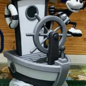 May include: Black and white figurine of Mickey Mouse as a steamboat captain, standing on a boat with a wheel and a life preserver. The base of the figurine is a film reel with images of Mickey Mouse.
