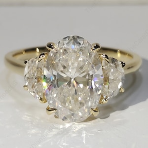 May include: A gold ring featuring a large, oval-cut diamond flanked by two smaller, oval-cut diamonds. The ring's band is a simple, polished gold. The diamonds sparkle brightly against a white background. The ring is a piece of fine jewelry.