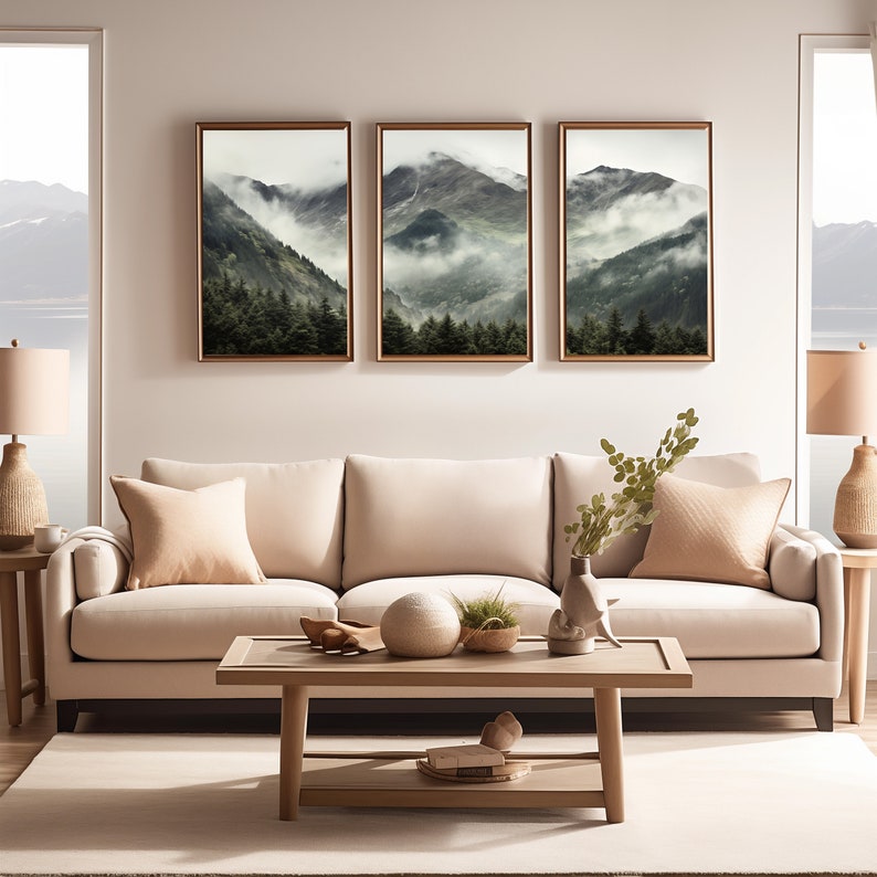 May include: Three framed landscape photographs of a mountain range with fog and trees. The photographs are hung on a light beige wall above a light beige sofa with two pillows. A coffee table with a vase of greenery, a round white decorative object, and a small basket of greenery sits in front of the sofa.
