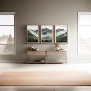 May include: Three framed landscape photographs of a mountain range with fog and trees, hanging on a light brown wall above a light wood console table with two drawers and a shelf. The console table is in front of a large window with a view of a mountain range.