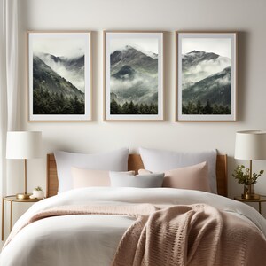 May include: Three framed landscape photographs of a mountain range with fog and trees. The photographs are hung on a white wall above a bed with white bedding and a pink throw blanket.