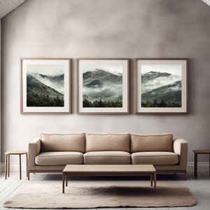 May include: Three framed landscape photographs of a mountain range with fog and trees. The photographs are hung on a light gray wall above a beige sofa and a wooden coffee table.