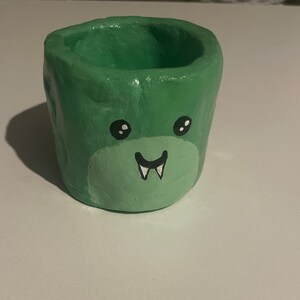 May include: A small green ceramic planter with a painted face, featuring black eyes, a white smile, and two fangs.