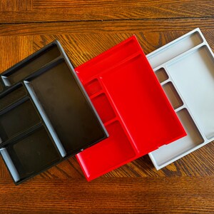 K&ouml;nnte beinhalten: Drei Plastik-Schreibtisch-Organizer mit verschiedenen F&auml;cherkonfigurationen. Die Organizer sind schwarz, rot und grau.