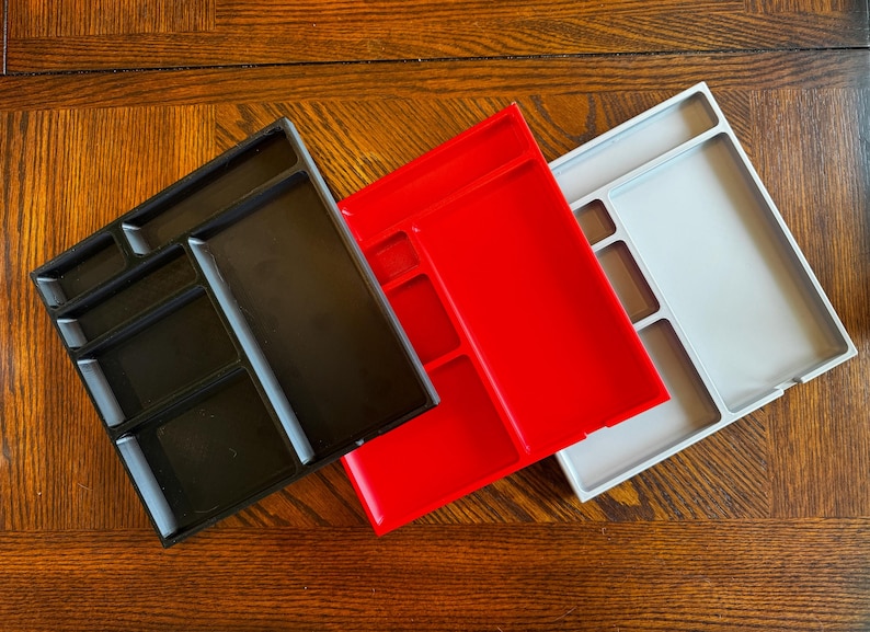 K&ouml;nnte beinhalten: Drei Plastik-Schreibtisch-Organizer mit verschiedenen F&auml;cherkonfigurationen. Die Organizer sind schwarz, rot und grau.