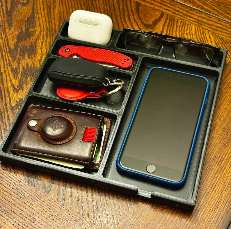 K&ouml;nnte beinhalten: Ein schwarzes Organizer-Tablett mit F&auml;chern f&uuml;r ein Telefon, eine Brieftasche, Schl&uuml;ssel und andere Kleinigkeiten. Das Tablett enth&auml;lt ein schwarzes Handy-Etui, eine braune Ledergeldb&ouml;rse, ein rotes Taschenmesser und ein wei&szlig;es Etui f&uuml;r kabellose Ohrh&ouml;rer.