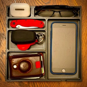 K&ouml;nnte beinhalten: Ein schwarzes Organizer-Tablett mit sechs F&auml;chern. Die F&auml;cher enthalten eine braune Ledergeldb&ouml;rse, einen schwarzen und silbernen Schl&uuml;sselanh&auml;nger mit einem roten Schl&uuml;sselanh&auml;nger, ein rotes Taschenmesser, ein wei&szlig;es kabelloses Ladeetui, eine schwarze Brille und ein schwarzes Smartphone.