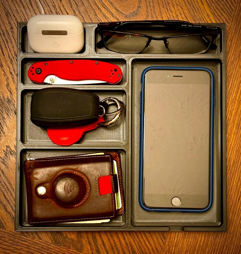 K&ouml;nnte beinhalten: Ein schwarzes Organizer-Tablett mit sechs F&auml;chern. Die F&auml;cher enthalten eine braune Ledergeldb&ouml;rse, einen schwarzen und silbernen Schl&uuml;sselanh&auml;nger mit einem roten Schl&uuml;sselanh&auml;nger, ein rotes Taschenmesser, ein wei&szlig;es kabelloses Ladeetui, eine schwarze Brille und ein schwarzes Smartphone.