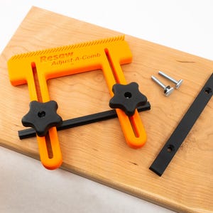May include: An orange plastic woodworking tool called "Resaw Adjust-A-Comb" with two black star-shaped knobs. The tool is on a wooden surface with two black metal strips and two silver screws.