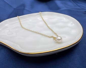 May include: A delicate gold chain necklace with a single white pearl pendant. The necklace is displayed on a white ceramic dish with a gold rim.